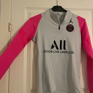 Paris Saint Germain  - Säljer en snygg vit psg tröja med rosa ärmar, tröjan har en halv dragkedja och ett Paris Saint-Germain-märke på bröstet. Perfekt för sportiga tillfällen eller som en stilren vardagströja.
