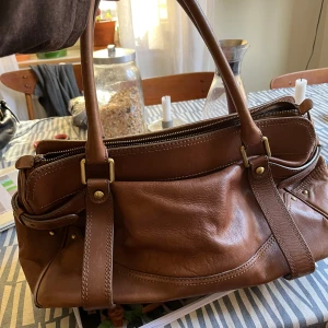 Brun skinnväska från Massimo Dutti - Säljer en snygg brun skinnväska med gulddetaljer från Massimo Dutti. Väskan har dubbla handtag och en praktisk dragkedja upptill. Perfekt för att bära dina dagliga nödvändigheter med stil. Inuti finns en etikett med märkesnamnet.