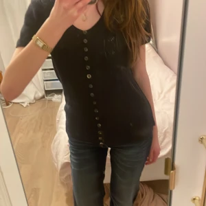 Svart kortärmad topp från KUS Stockholm - Säljer en stilren svart kortärmad topp från KUS Stockholm. Toppen har en v-ringning och knappar framtill, vilket ger en elegant touch. Perfekt för att matcha med jeans eller kjol. Passar bra för både vardag och speciella tillfällen.