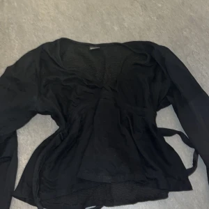 Svart omlottopp från Gina Tricot - Säljer en svart Texturerad topp med knytning bak. Toppen är vit och har v-ringning. Den har normal passform, utsvängd ärm och rynkningar under bysten från Gina Tricot. 