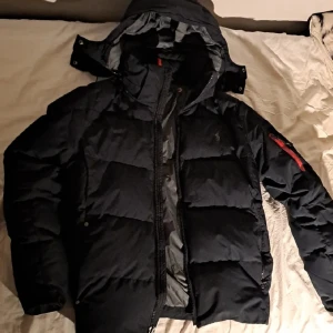 Jacka från Polo Ralph Lauren - Mörkblå dunjacka från Polo Ralph Lauren med en cool röd detalj på ärmen. Jackan har en huva och dragkedja framtill. (Min sons jacka, som är ca 180 cm.)
