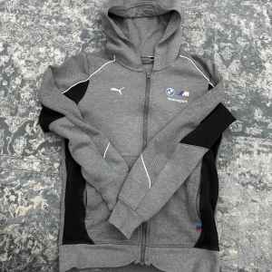Grå hoodie från Puma - Säljer en grå hoodie från Puma med BMW Motorsport-logga. Tröjan har dragkedja och svarta detaljer på sidorna. Perfekt för bilentusiaster!