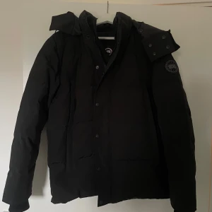 Svart dunjacka från Canada Goose (black label) - Säljer en svart dunjacka från Canada Goose med lång ärm och huva. Jackan har en broderad logotyp på ärmen och stängs med knappar och dragkedja. Perfekt för kalla vinterdagar.