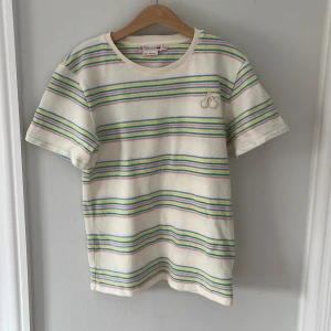 Randig t-shirt från Bonpoint - Säljer en söt randig t-shirt från Bonpoit med ett körsbär på bröstet. T-shirten har korta ärmar och är i mjukt material med färgglada ränder i blått, gult, rosa och grönt på en vit bas. Frakt ingår inte i priset!