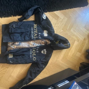 Svart jacka från SANTOOZ ROYAL POLO - Snygg svart jacka med huva från SANTOOZ ROYAL POLO. Jackan har flera tryck i guld och vitt, samt en stor logga på ryggen. Insidan är mönstrad med repmotiv i brunt och beige. Perfekt för en stilren look.