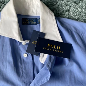 Blå skjortklänning från Polo Ralph Lauren - Elegant blå skjortklänning från Polo Ralph Lauren med vit krage och knappar framtill. Klänningen har långa ärmar och en klassisk passform som ger en stilren look.
