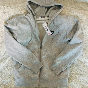 Grå hoodie från Comme des Garçons - Säljer en stilren och exklusiv zip-hoodie från Comme des Garçons i grått, med snygga tryck både på bröstet och ryggen. Perfekt för dig som vill ha en hoodie som sticker ut men ändå är lätt att matcha med olika outfits!   Skick: Helt ny och aldrig använd – endast provad. Storlek:  M  Fick den i födelsedagspresent, men den var tyvärr  för stor för mig.  Hör av dig om du har frågor!