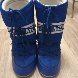 Blå Moon Boots - Säljer ett par klassiska blå Moon Boots med vit logga och snörning. Perfekta för vintern med sin varma och vadderade design. De har en rund tå och en robust sula för bra grepp på snöiga underlag. Se bild 3, slitage nere på ena skon