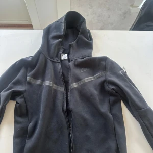 Svart hoodie från Nike - Säljer en svart hoodie från Nike med dragkedja och huva. Jackan har en diskret logga på bröstet och detaljerade sömmar. Perfekt för en sportig look.