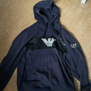 Mörkblå hoodie från Emporio Armani - Snygg mörkblå hoodie från Emporio Armani med det ikoniska EA7-logotypen på bröstet och ärmen. Hoodien har en klassisk passform med justerbar huva och ribbade muddar. Passar perfekt i storlek L och XL.