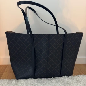 Malene Birger väska - Snygg och rymlig svart shoppingväska från malene birger med ett diskret rutigt mönster. Väskan har långa axelremmar som gör den bekväm att bära. Perfekt för att bära med sig allt man behöver under dagen.