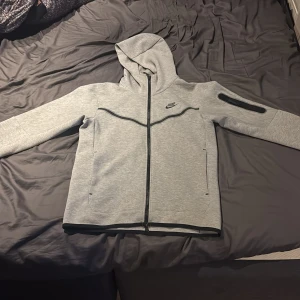 Grå Nike Tech hoodie - Säljer en stilren Nike Tech från Nike knappt använd i fint skick med dragkedja och huva. Jackan har en ficka med dragkedja på ärmen och två sidofickor. 