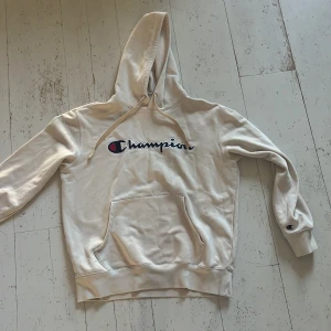 Vit hoodie från Champion - Säljer en vit hoodie från Champion med klassisk logga på bröstet. Tröjan har en stor ficka framtill och justerbar huva med dragsko. Tillverkad i en mjuk bomullsblandning för extra komfort.