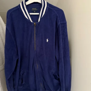 Blå kofta från Polo Ralph Lauren - Säljer en snygg blå kofta från Polo Ralph Lauren. Koftan har en dragkedja framtill och vita ränder på kragen. Den ikoniska loggan är broderad på bröstet. Perfekt för en stilren look!