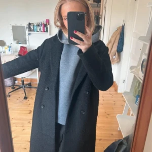 Svart kappa - Snygg svart kappa med dubbelknäppning och klassisk design. Perfekt för kyligare dagar med sin långa passform och stilrena look. Passar bra över en hoodie för en avslappnad stil.