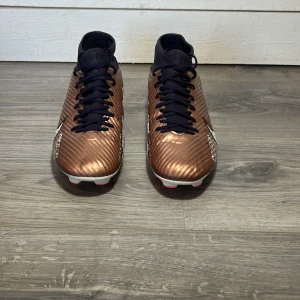 Nike Mercurial fotbollsskor i brons och svart - Snygga Nike Mercurial fotbollsskor i en cool bronsfärg med svarta detaljer. Skorna har snörning och en hög ankelkrage för extra stöd. Perfekta för fotbollsplanen med sin avancerade design och greppvänliga dobbar.