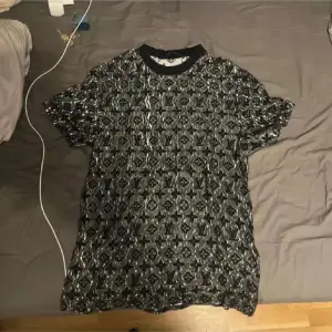 Säljer en snygg t-shirt från Louis Vuitton med ett unikt svart och grått mönster. T-shirten är kortärmad och tillverkad i Italien. Perfekt för dig som vill ha något stilrent och exklusivt.