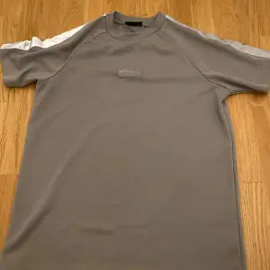 Snygg grå t-shirt från McKenzie med korta ärmar och vita detaljer längs axlarna. Perfekt för en avslappnad stil. Tillverkad i ett mjukt syntetmaterial för extra komfort.