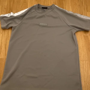 Grå t-shirt från McKenzie - Snygg grå t-shirt från McKenzie med korta ärmar och vita detaljer längs axlarna. Perfekt för en avslappnad stil. Tillverkad i ett mjukt syntetmaterial för extra komfort.