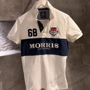 Vit och marinblå pikétröja från Morris - Snygg vit pikétröja från Morris med marinblått band och broderad logga. Tröjan har korta ärmar och knappar vid kragen. Perfekt för en stilren look.