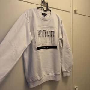 Vit sweatshirt från SMOG - Säljer en vit sweatshirt från SMOG med texten 'ICONO MILANO' i svart och vitt på framsidan. Tröjan har en normal passform och är tillverkad i ett mjukt material som känns skönt mot huden. Perfekt för en avslappnad stil.