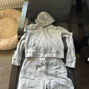 Grå hoodie och byxor från Juicy Couture - Säljer en grå hoodie och matchande byxor från Juicy Couture. Hoodien har dragkedja och snörning i luvan. Byxorna har praktiska fickor med knappar. Perfekt för en avslappnad stil. 🩶