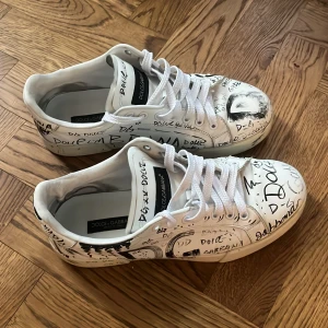 Vita sneakers med svart graffiti från Dolce & Gabbana - Unika vita sneakers från Dolce & Gabbana med handskriven graffiti-design i svart. Skorna har snörning och en rund tå, dessa är dem perfekta sommar skorna, som sticker ut lagom men passar till de mesta.