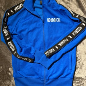 Blå set från Hoodrich - Snygg blå träningsjacka från Hoodrich med dragkedja och logotyp längs ärmarna. Jackan har en normal passform och är perfekt för en sportig look. Den är tillverkad i ett mjukt syntetmaterial och har ribbade muddar för extra komfort. Det kommer med byxorna alltså hela setet, Kan köpas separat om önskas det är bara att skriva Storlek S