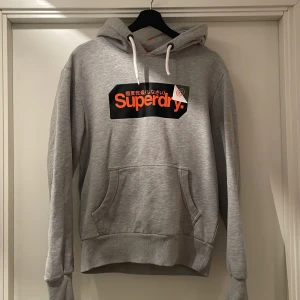 Grå hoodie från Superdry - Säljer en grå hoodie från Superdry med en stor logga i orange och svart på bröstet. Tröjan har en klassisk känguruficka och justerbar huva med vita snören. Perfekt för en avslappnad stil.