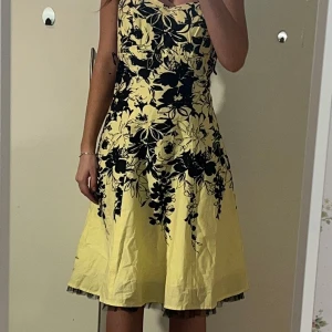 Gul blommig klänning - Säljer en söt gul klänning med svart blommönster. Klänningen har en v-ringad design och är ärmlös, vilket gör den perfekt för varmare dagar. Den har en bekväm passform och når till knäna. Perfekt för sommarens alla tillfällen!
