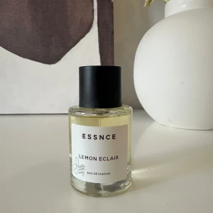 Essnce Lemon Eclair Eau de Parfum - Fräsch och uppfriskande parfym från Essnce med doft av citron och eclair. Flaskan är stilren och genomskinlig med en svart kork och vit etikett. Perfekt för dig som älskar en söt och citrusaktig doft.
