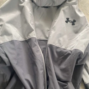 Grå vindjacka från Under Armour - Säljer en stilren grå vindjacka från Under Armour med dragkedja framtill. Jackan har en hög krage och det ikoniska logotypen på bröstet. Perfekt för träning eller en promenad i blåsigt väder.