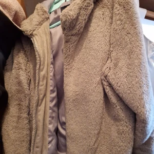 Beige teddyjacka från Vero Moda - Mysig beige teddyjacka från Vero Moda med dragkedja framtill. Perfekt för kyliga dagar när du vill hålla dig varm och stilren. Jackan har en mjuk och fluffig yta som ger en trendig look.