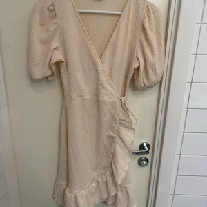 Beige omlottklänning - Säljer en söt beige omlottklänning med volangdetaljer. Klänningen har korta puffärmar och en v-ringad design. Perfekt för sommarens alla tillfällen. 💫