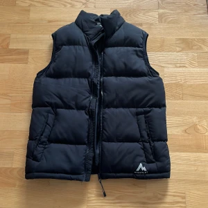 Svart dunväst från McKinley - Säljer en svart dunväst från McKinley med dragkedja och praktiska fickor. Perfekt för kyliga och även varmare dagar. Har en stilren design med logotypen på framsidan. Passar bra för utomhusaktiviteter.