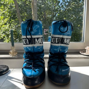 Blå Moon Boot snowboots - Säljer ett par ikoniska Moon Boot snowboots i blått och mörkblått med vit logga runt skaftet. Skorna har snörning framtill och är tillverkade i syntetmaterial med platt sula. Perfekta för vinterns kalla dagar!! 