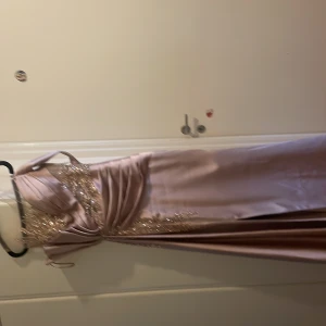 Champagnefärgad långklänning med paljetter - Säljer en elegant champagnefärgad långklänning med glittriga paljettdetaljer och broderier över livet. Klänningen har en axelbandslös design med transparent mesh upptill och vackra draperingar i midjan. Perfekt för bal eller nyår! Materialet är satin och mesh.