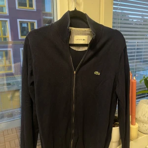 Mörkblå full zip från Lacoste - Snygg mörkblå full zip från Lacoste. Storlek M. Flärpen på zippern har ramlat av, annars fin. 