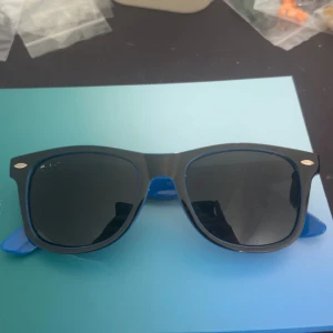 Blå och svarta Ray-Ban Wayfarer - Snygga Ray-Ban Wayfarer solglasögon med blå bågar och svarta skalmar. Glasen är mörka och har en klassisk fyrkantig form. Perfekt för dig som vill sticka ut med en cool färgkombo och ikonisk design. Ray-Ban-loggan i silver på sidan ger extra stilpoäng.