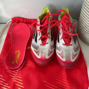 Adidas F50 fotbollsskor i vitt och rött - Säljer ett par Adidas F50 fotbollsskor i vitt med röda och svarta detaljer samt gula inslag. Skorna har snörning och en sportig, aerodynamisk design. Ovandelen är i syntetmaterial och sulan är tunn och smidig för bästa grepp på planen. Perfekta för dig som vill sticka ut på matchen. Använda få tal gånger och fungerar helt perfekt 