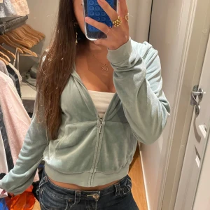 Ljusgrön velourhoodie Juicy Couture - Supermjuk ljusgrå hoodie från Juicy Couture i velour med dragkedja och klassisk huva. Croppad modell med långa ärmar och två fickor framtill. Ikonisk J-dragkedja i silverfärg. Perfekt för en chill och trendig look. Alldrig använd!!!