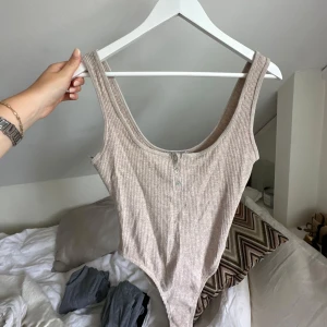 Beige ribbad body med knappar - Supersnygg beige ribbad body med djup urringning och små knappar framtill. Perfekt till jeans eller kjol för en chill och trendig look. Mjuk och stretchig, sitter tajt och bekvämt.