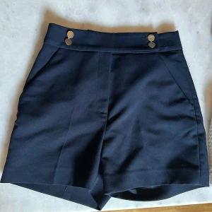 Marinblåa högmidjade shorts från H&M - Snygga marinblåa shorts från H&M i storlek 34. De är högmidjade med två guldiga knappar framtill. 