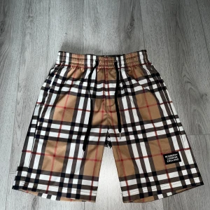 Burberry shorts  - Snygga Burberry shorts med klassiskt rutigt mönster i beige, svart, vitt och rött. Modellen har elastisk midja med svart snörning och en patch med Burberry London England på benet. Perfekt för dig som vill sticka ut med ikonisk design och skön passform.