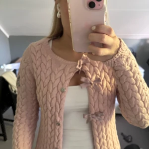 Rosa kofta från Odd Molly med rosetter - Superfin ljusrosa kofta från Odd Molly med grovstickat flätmönster och söta rosetter framtill. Koftan har lång ärm, rund hals och dekorativt Odd Molly-broderi på ena ärmen. Perfekt för dig som gillar gulliga detaljer och mjuka material.