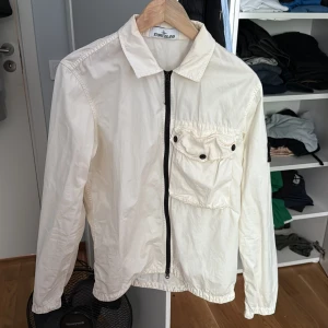 Vit Cardigan från Stone Island M - Säljer en clean vit vindjacka från Stone Island i storlek M. Jackan har dragkedja framtill, stor bröstficka med knappar och den klassiska Stone Island-patchen på ärmen. Perfekt för dig som gillar stilren streetwear med premiumkänsla.