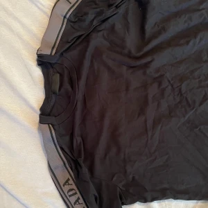 Svart t-shirt med gråa axelband - Svart t-shirt från Prada med gråa band längs axlarna där märkesnamnet syns. Klassisk rund halsringning och korta ärmar. Storlek M