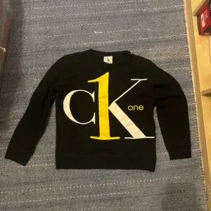 Svart Calvin Klein tröja med stor logga - Svart långärmad tröja från Calvin Klein Jeans med stor CK one-logga i vitt och gult framtill. Tröjan har rund halsringning och ribbade muddar. Perfekt för dig som gillar streetstyle och tydliga loggor.