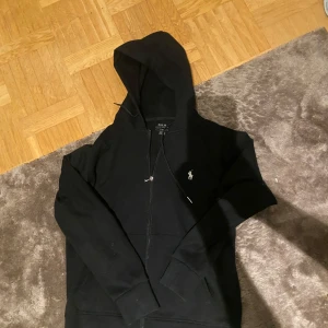 Svart hoodie från Ralph Lauren - Svart hoodie från Ralph Lauren med klassisk vit broderad logga på bröstet. Tröjan har dragkedja framtill, huva med snörning och två fickor. Tillverkad i mjuk bomull, perfekt för chill dagar. Långärmad och stilren design som funkar året runt. Självklart äkta👊Skick är 8/10 är en Size S några frågor är det bara att ställa🧐
