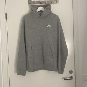 Grå hoodie från Nike med logga - Snygg grå hoodie från Nike med vit broderad logga på bröstet. Klassisk känguruficka framtill och huva. Perfekt för chill dagar eller när du vill ha en enkel och stilren look. Mjuk insida och normal passform.